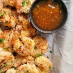 coconut shrimp garnished with fresh chopped parsley on a parchment lined baking sheet with an orange dipping sauce
