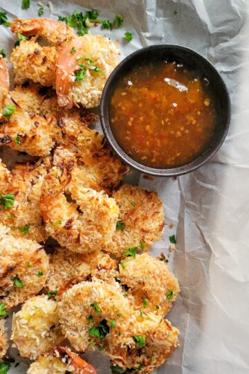 coconut shrimp garnished with fresh chopped parsley on a parchment lined baking sheet with an orange dipping sauce
