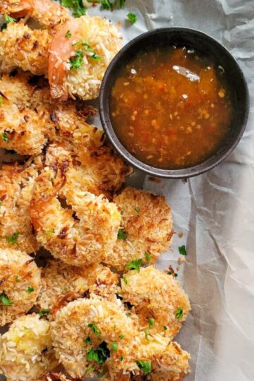 coconut shrimp garnished with fresh chopped parsley on a parchment lined baking sheet with an orange dipping sauce