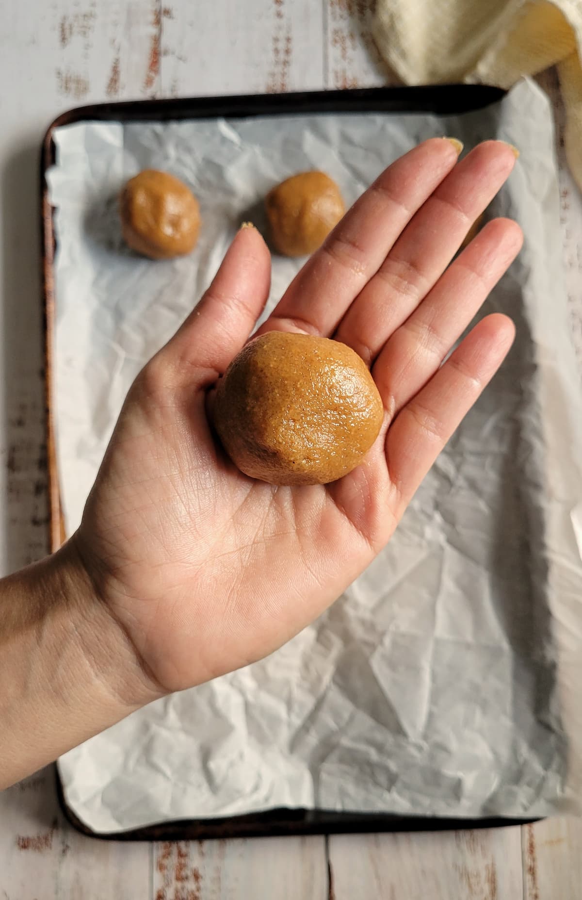 ball of cookie dough in the palm of a hand over a parchment lined baking sheet with 2 more dough balls