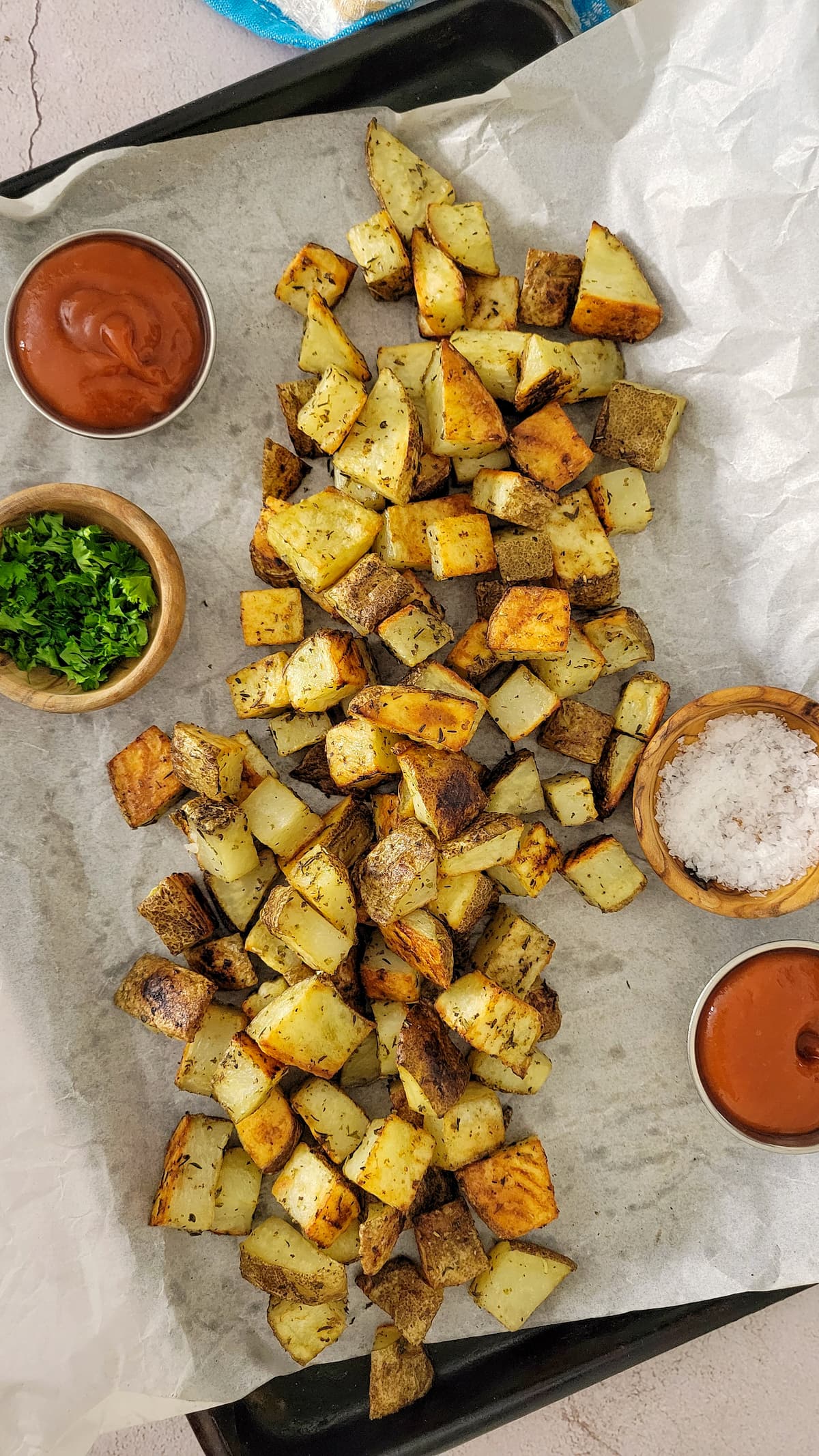 pile of crispy home fries on a parchment lined baking sheet, ramekins of salt, ketchup and parsley around it