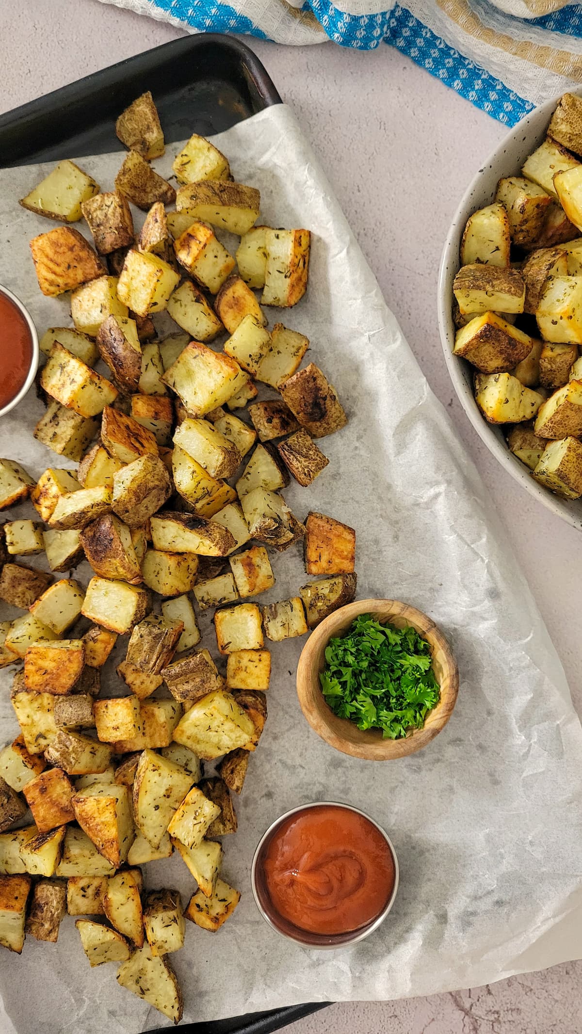 crispy home fries on a parchment lined baking sheet with ramekins of ketchup and parsley, bowl of more potatoes next to it