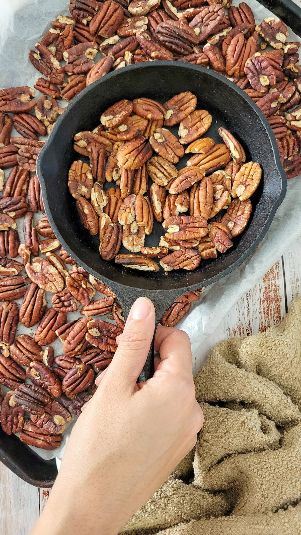 hand holding a skillet with toasted pecans on top of a parchment lined baking sheet with more