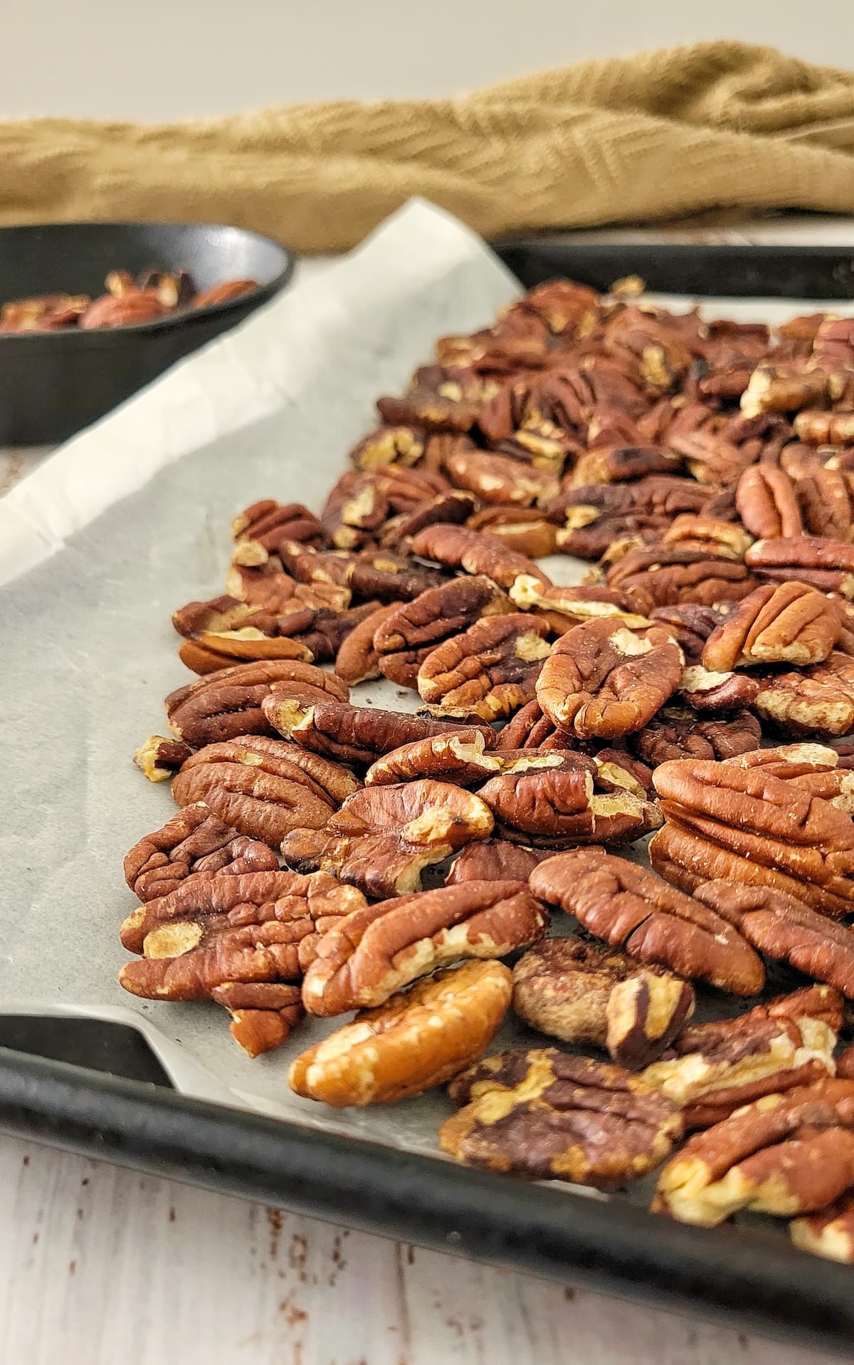 side view of toasted pecans on a parchment lined baking sheet, more in a skillet in the background
