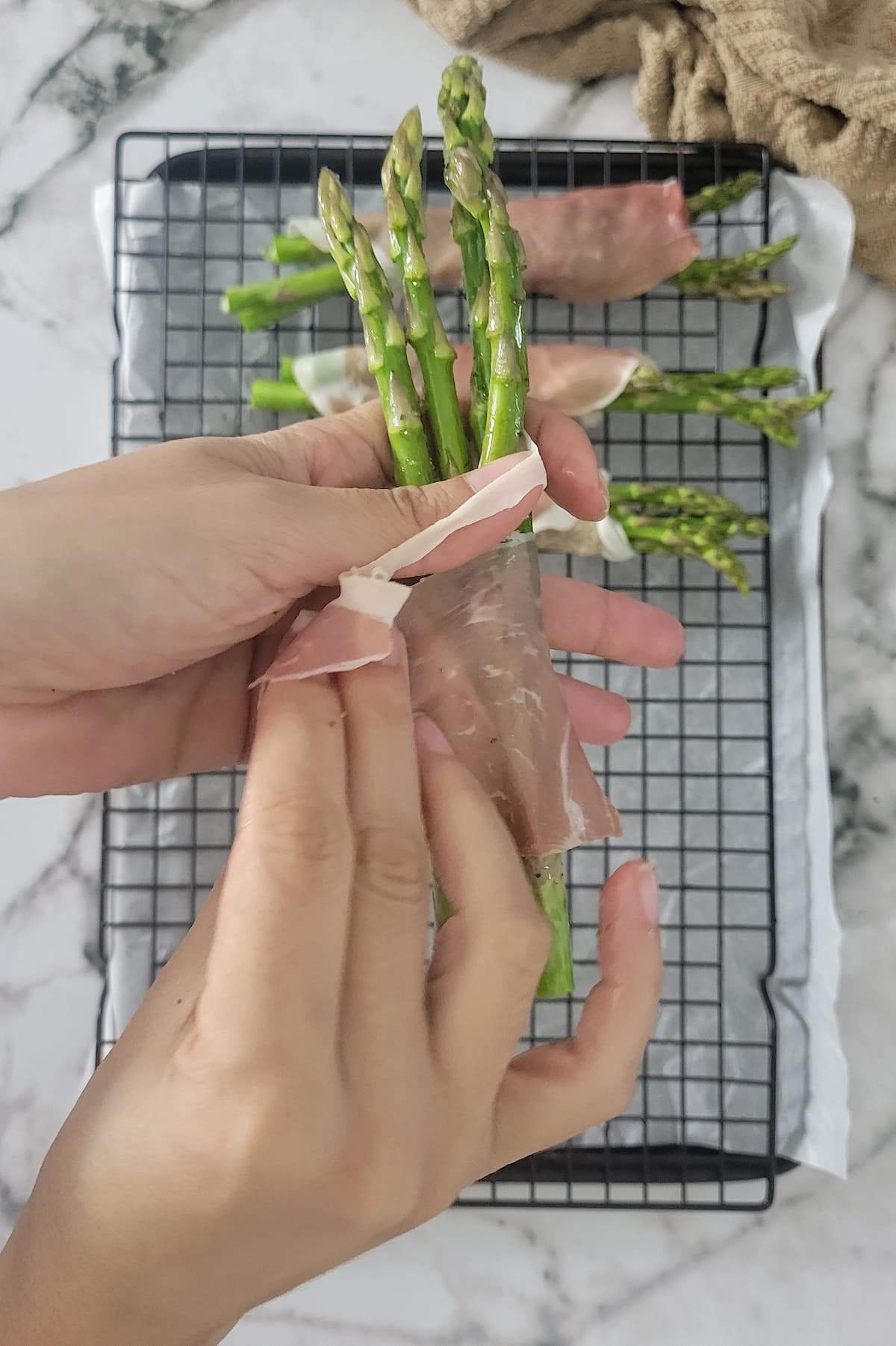 hands wrapping a piece of prosciutto around a bundle of asparagus over a tray with 3 more bundles