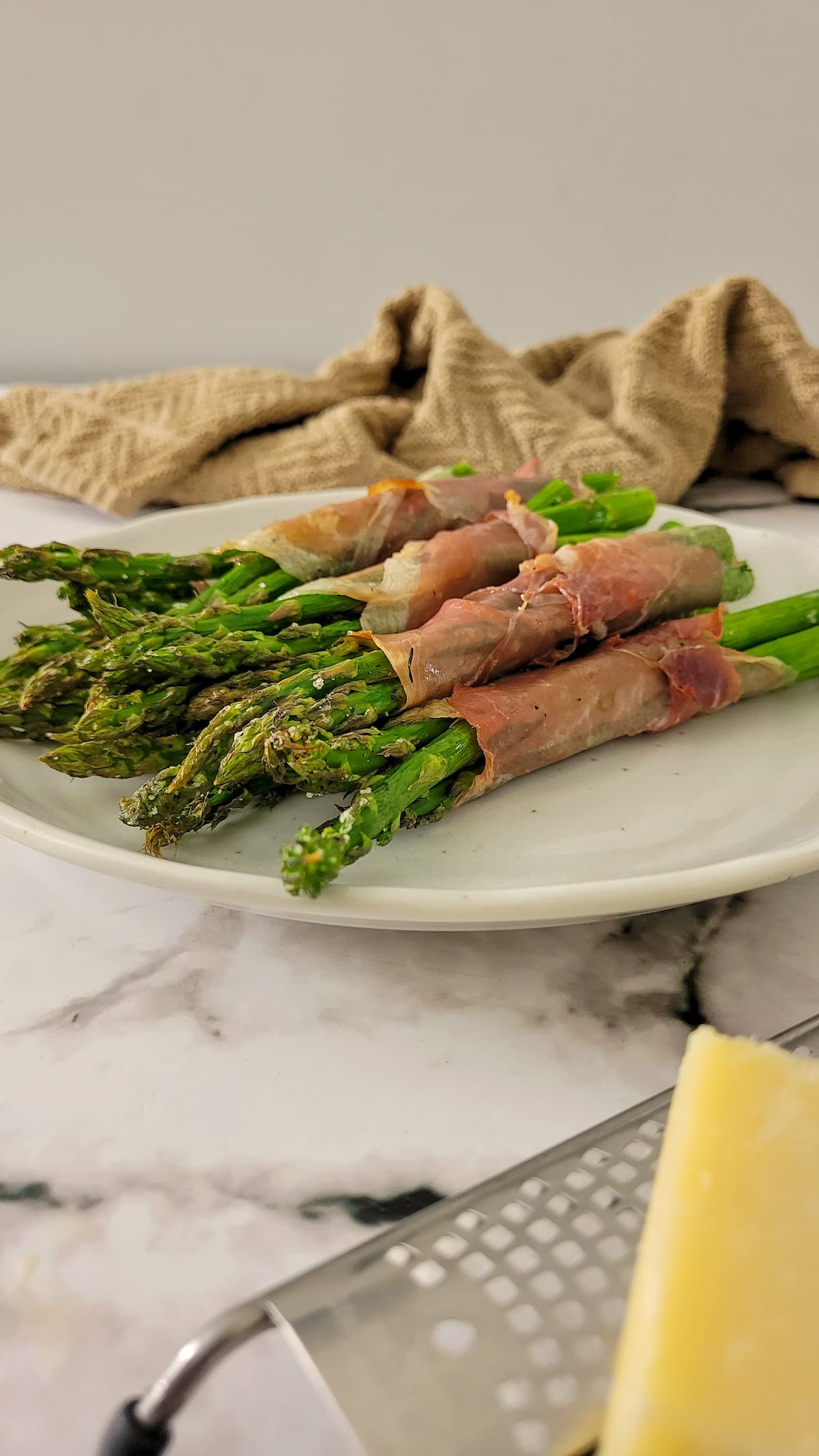 side view of prosciutto wrapped asparagus bundles on a plate, grater with parmesan in the front