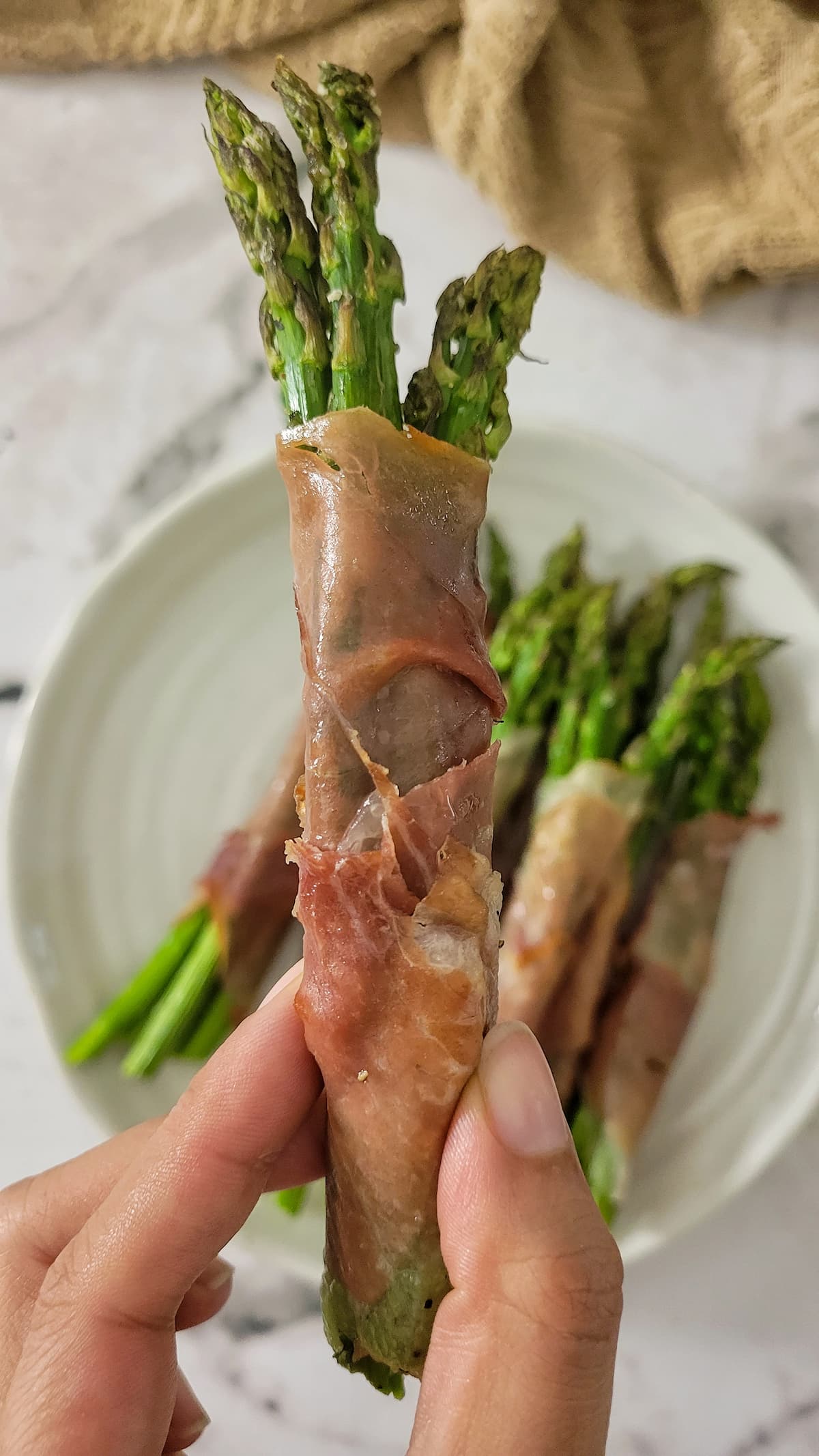 hand holding a bundle of asparagus wrapped with prosciutto over a plate with the rest of them