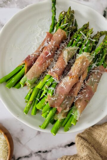 prosciutto wrapped asparagus bundles on a plate garnished with fresh grated parmesan cheese