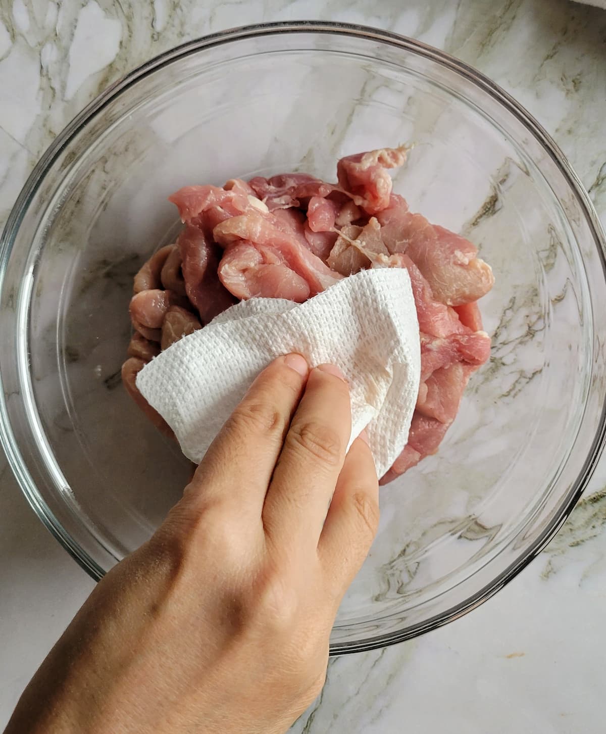hand with a paper towel patting down raw pork strips in a bowl