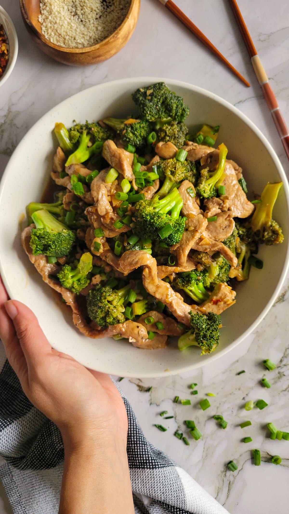 hand holding a bowl of stir fried broccoli and pork garnished with green onions