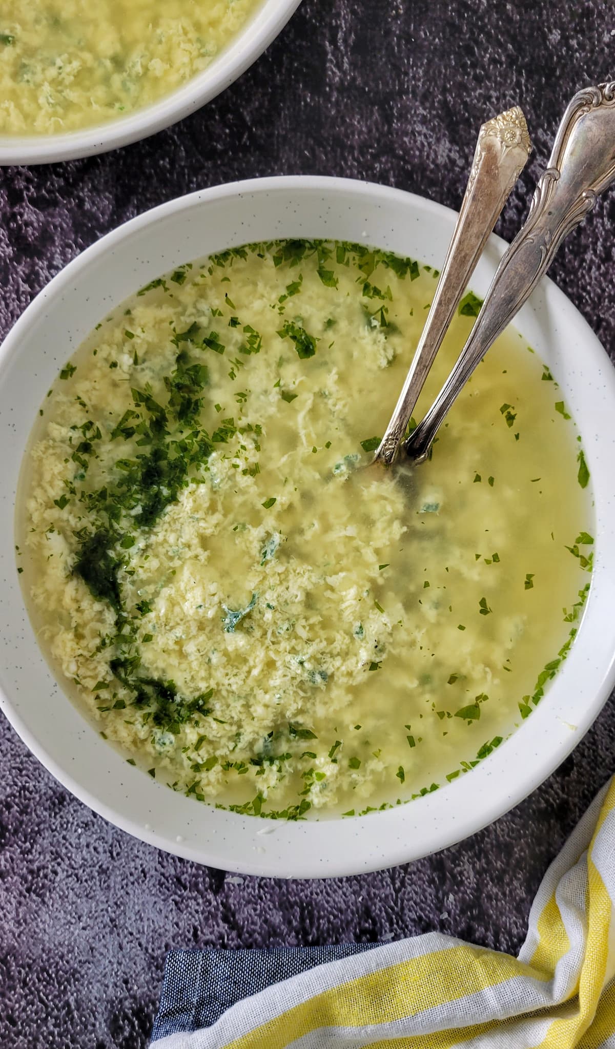 bowl of egg drop soup with chopped fresh parsley, two spoons in the bowl, another bowl of soup in the background