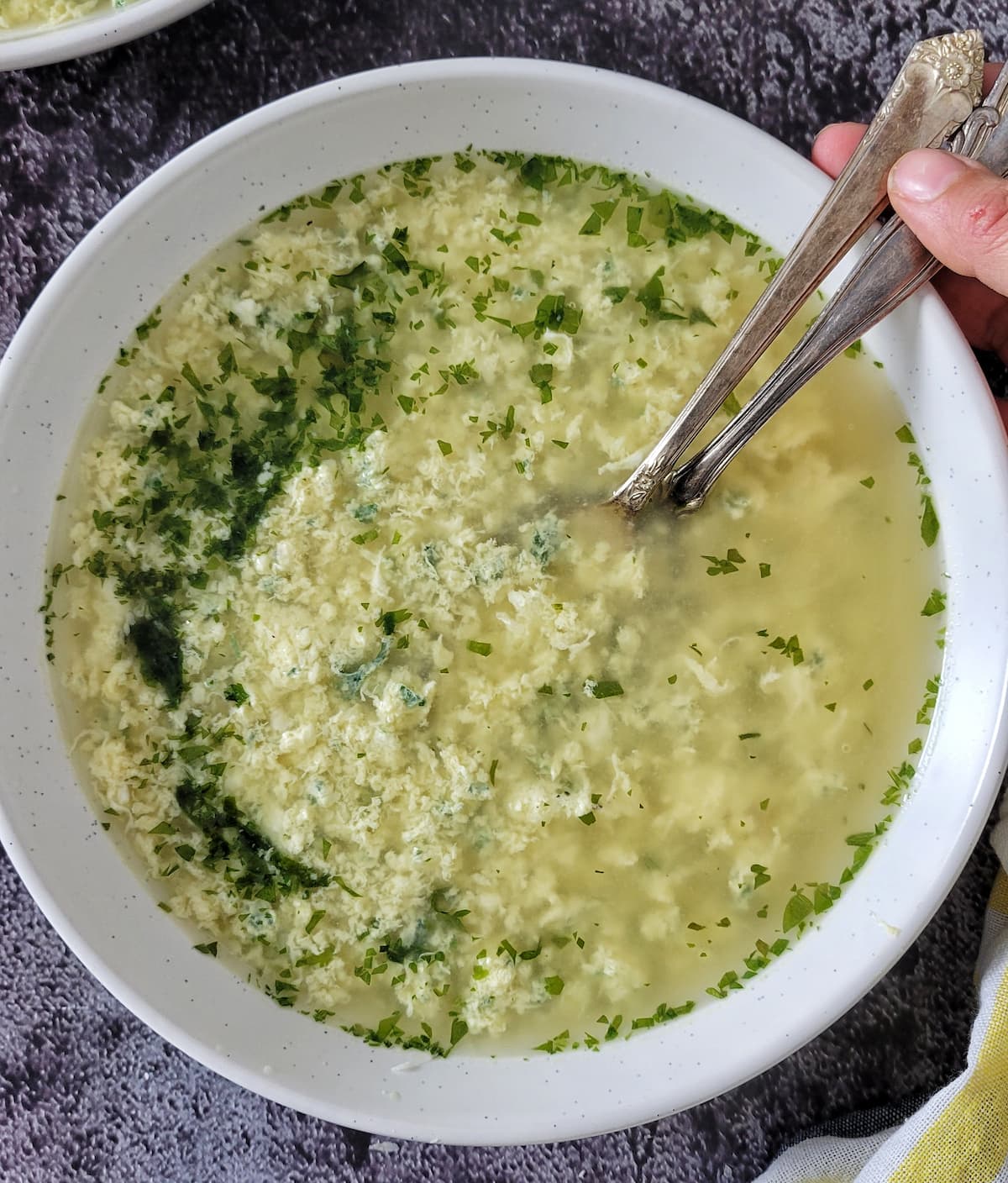 hand holding two spoons in a bowl of egg drop soup with parsley