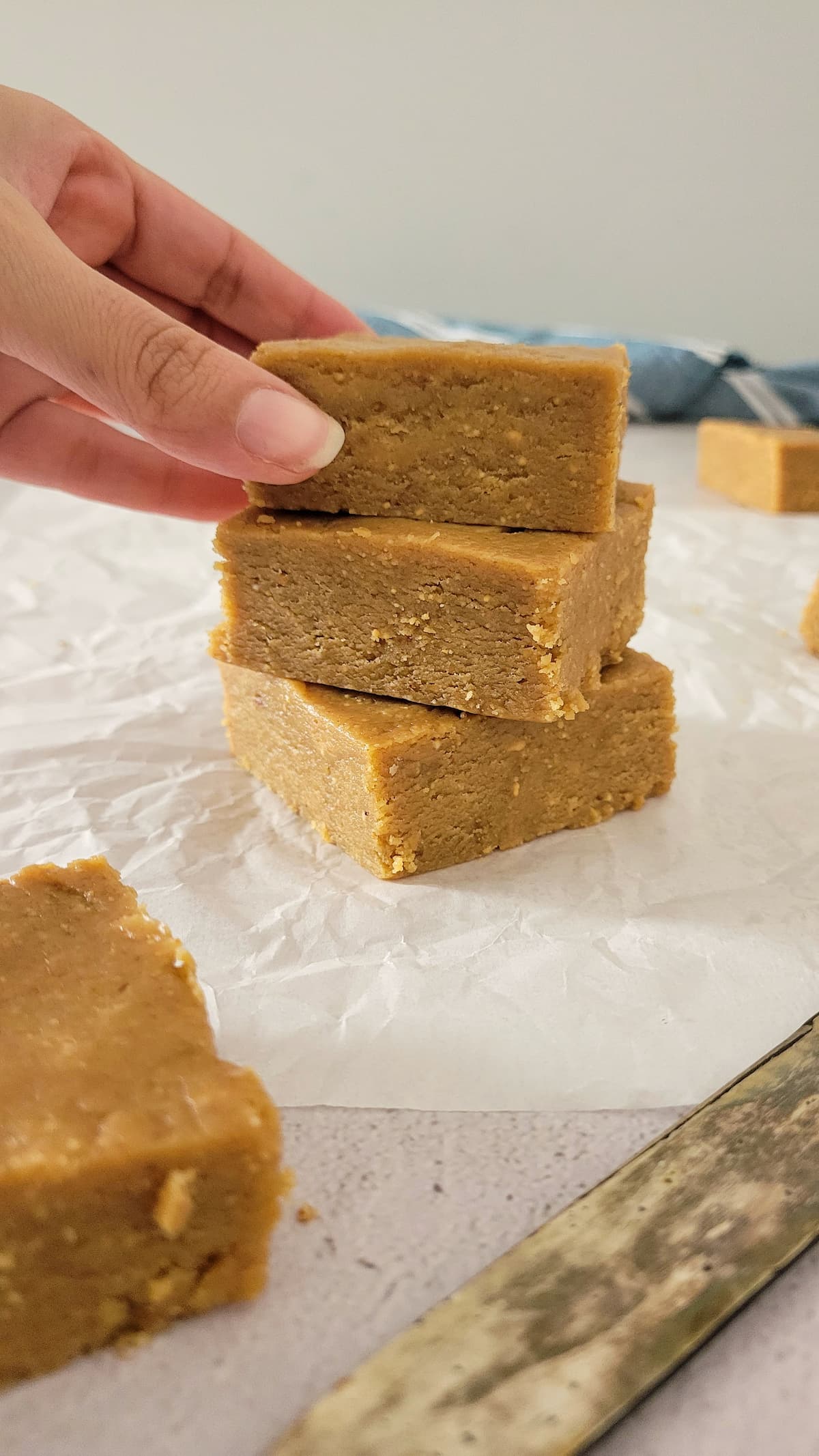 3 pieces of peanut butter fudge stacked onto a piece of parchment paper, more fudge in the background, hand grabbing the first piece