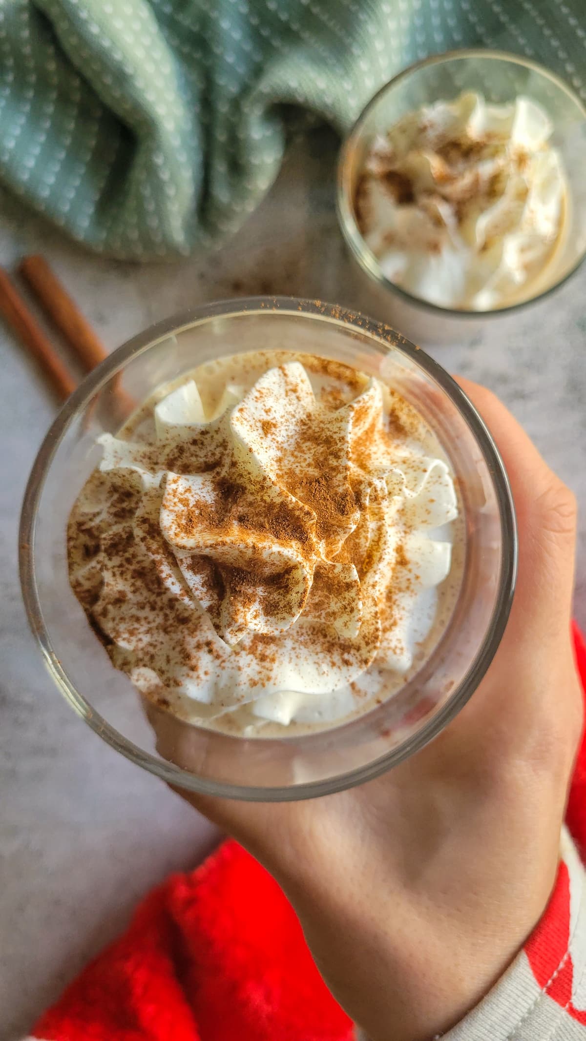 hand holding a glass of eggnog with whipped cream and cinnamon over another glass with the same thing