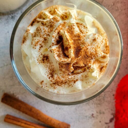 eggnog with whipped cream and cinnamon next to two cinnamon sticks