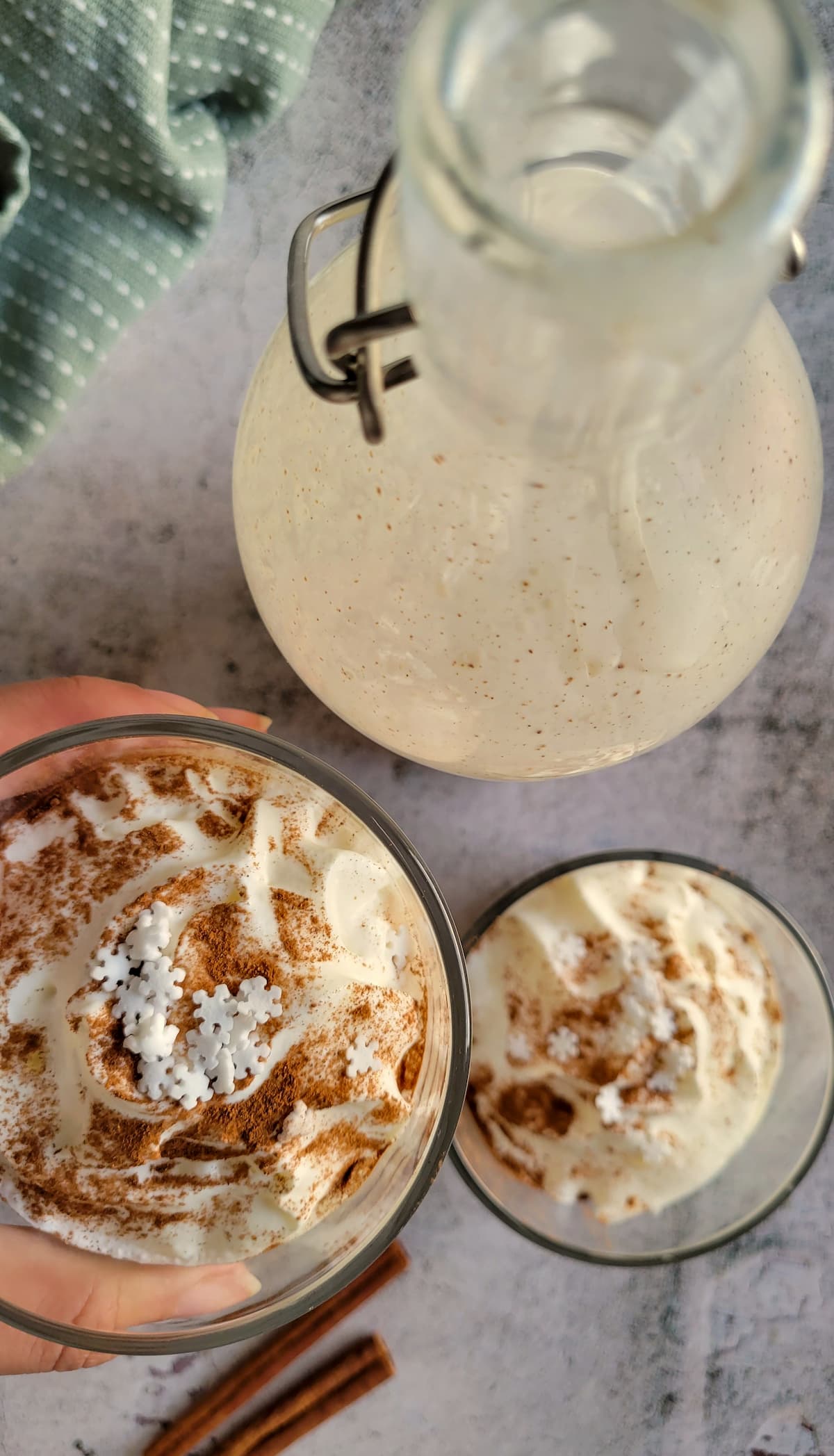 two glasses of eggnog topped with whipped cream, cinnamon and snowflake sprinkles next to a jug of eggnog and cinnamon sticks