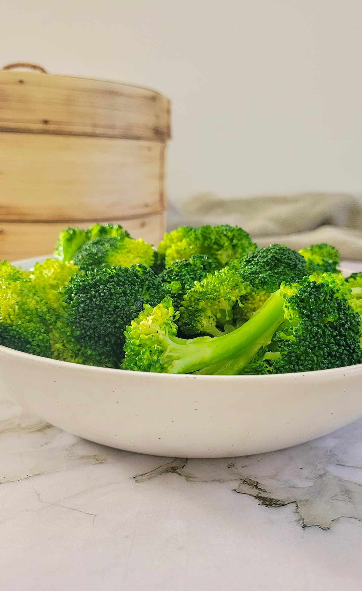 side view of a bowl of broccoli, bamboo steamer set in the background