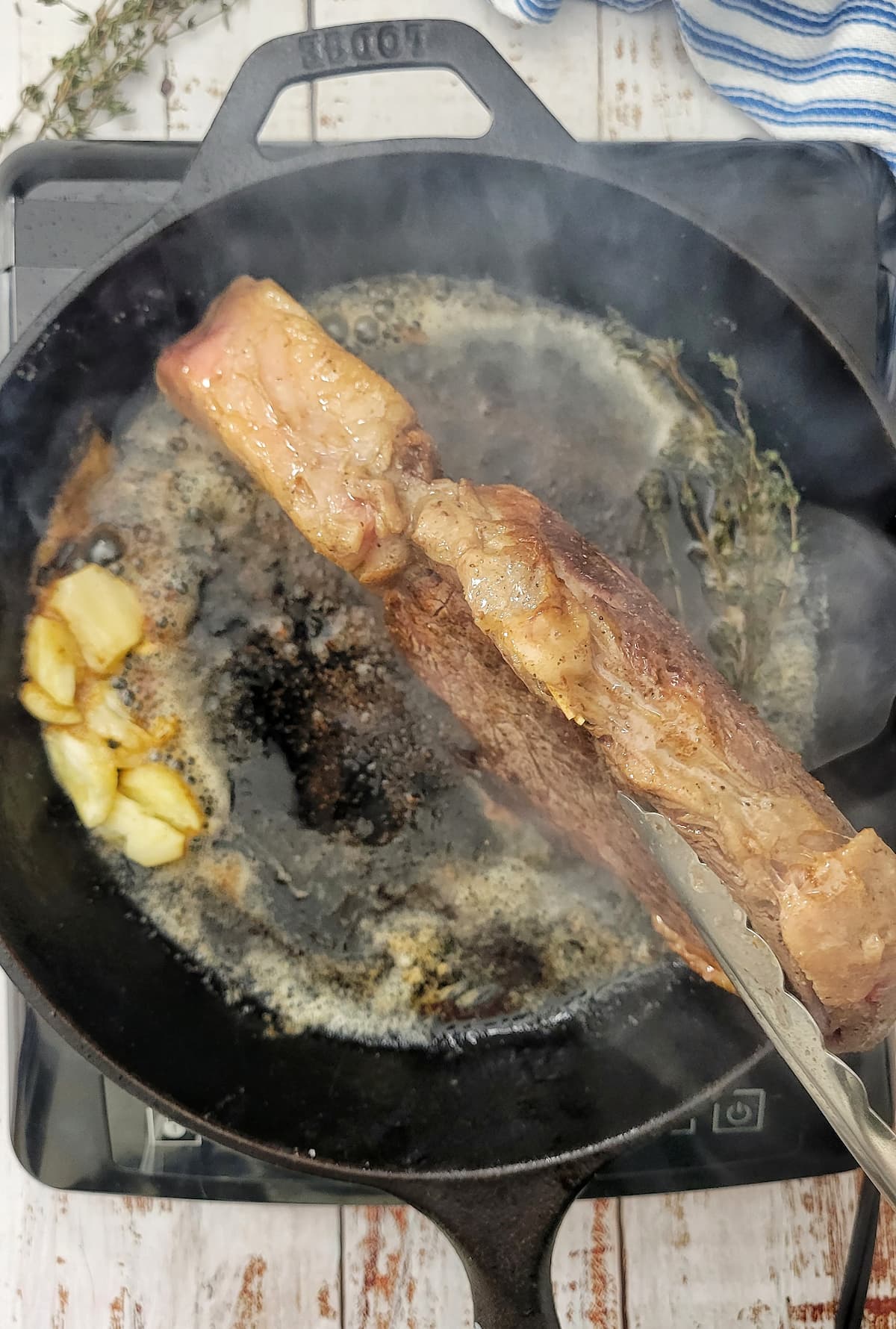 tongs holding a steak on it's side to cook in a cast iron skillet with fresh herbs and garlic