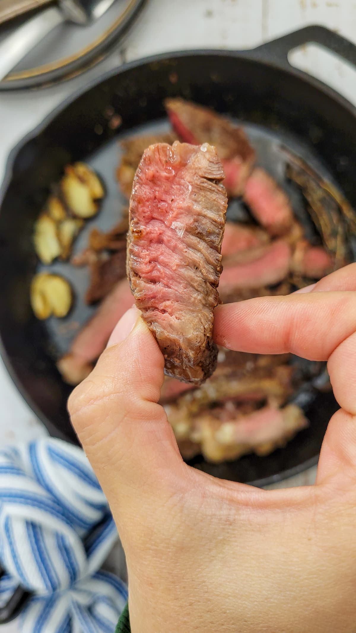 hand holding a piece of medium rare steak over a skillet with the rest of it