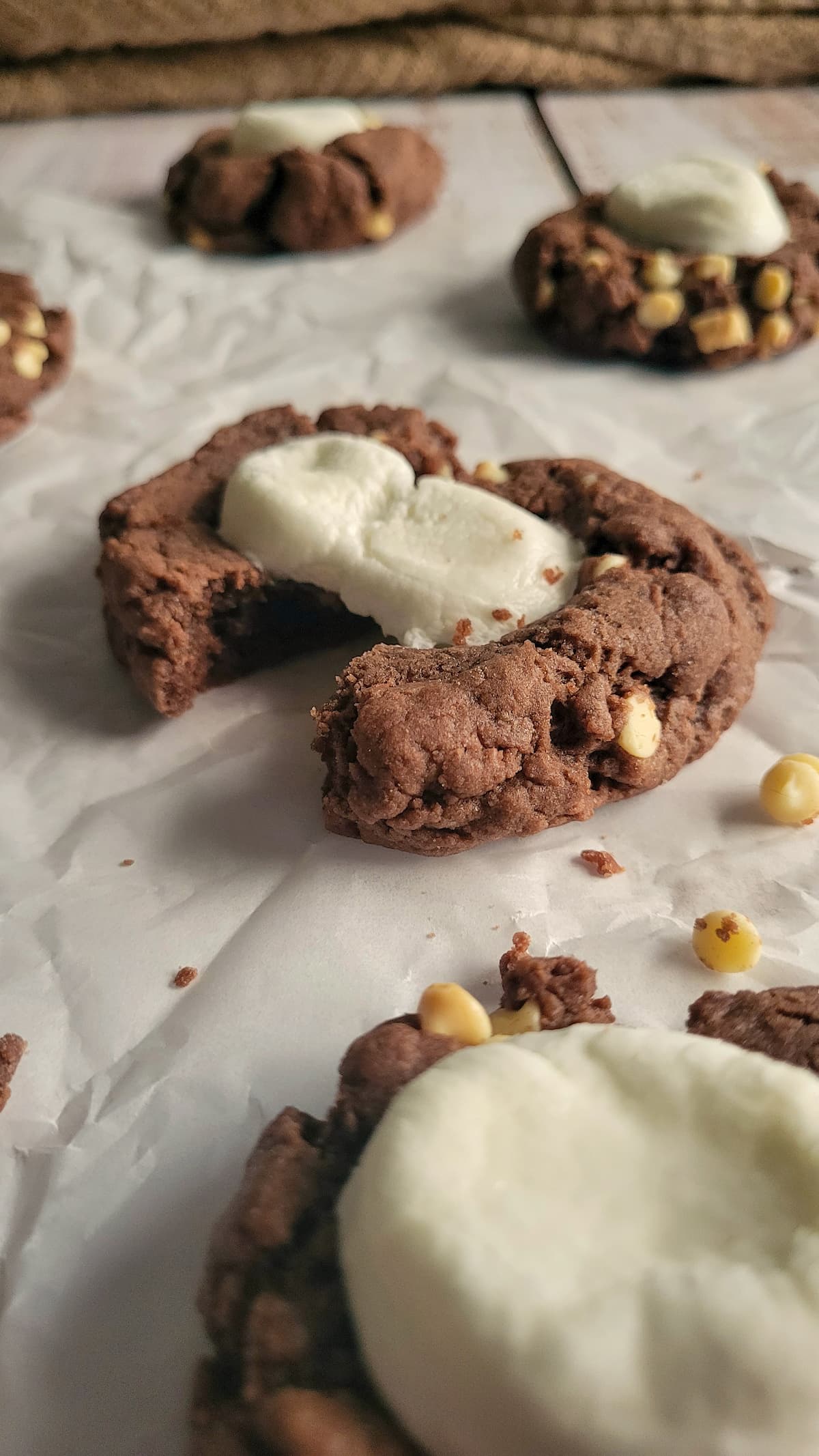 hot cocoa cookies with white chocolate chips and a giant marshmallow on a piece of parchment paper, first cookie broken in half
