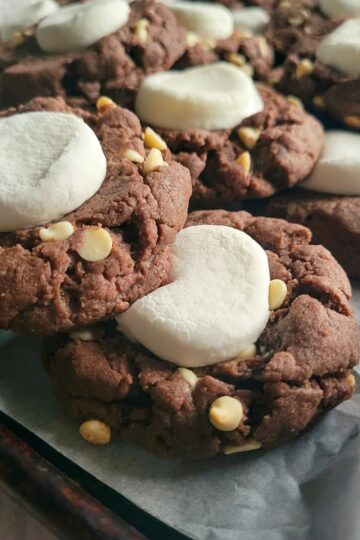 hot cocoa cookies with white chocolate chips and marshmallow stacked on a parchment lined baking sheet