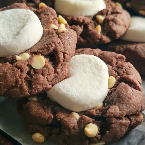chocolate cookies with white chocolate chips and marshmallow stacked on a parchment lined baking sheet