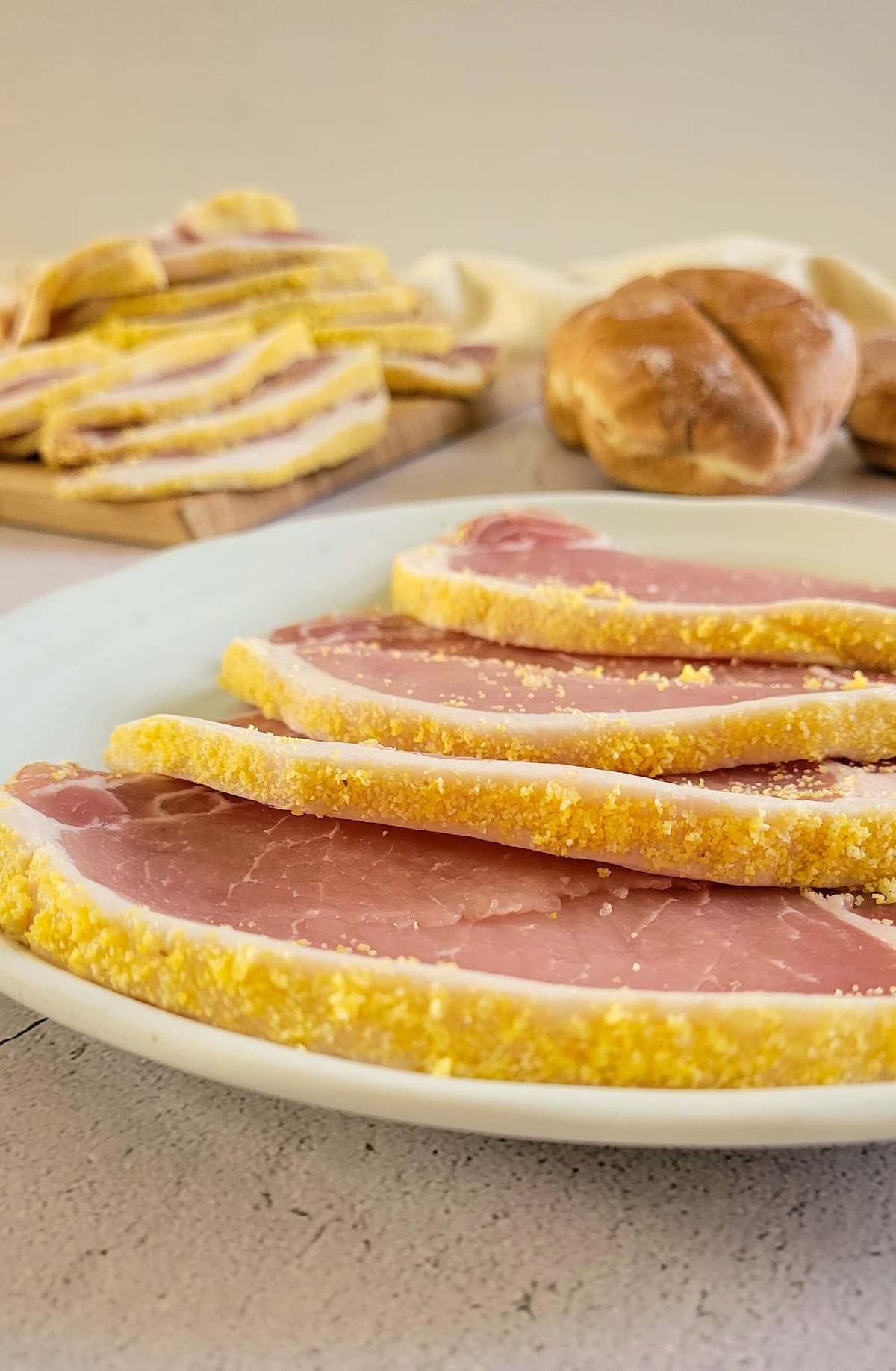 raw slices of peameal bacon on a plate, more in the background with buns