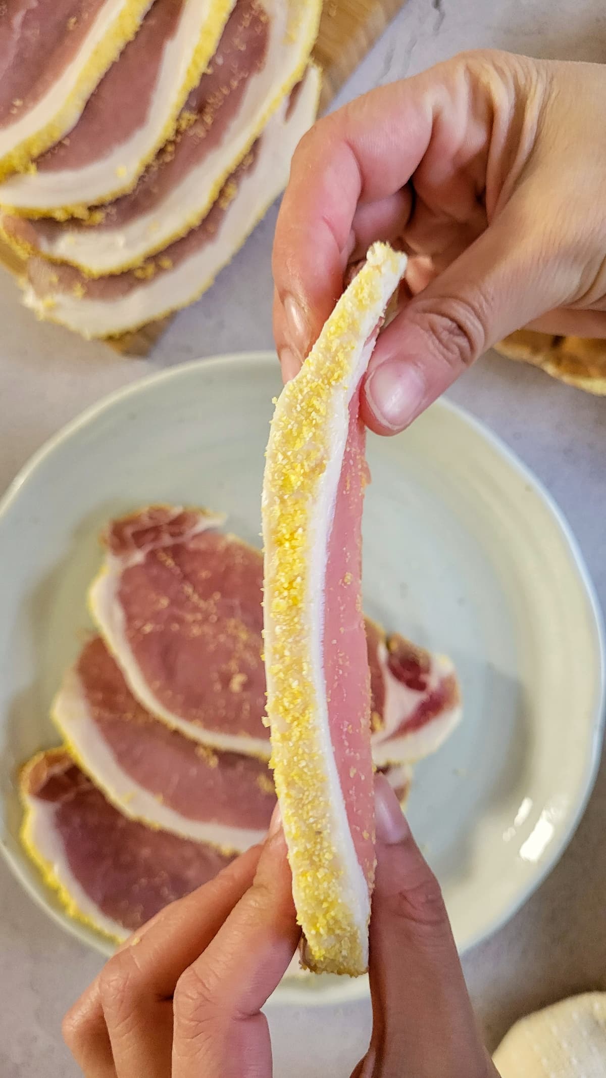 hand holding a raw slice of peameal bacon on its side over a plate with more