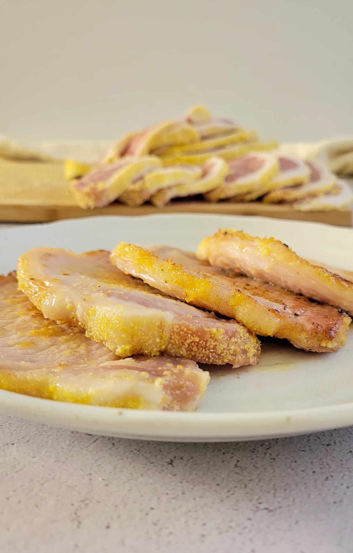 4 slices of cooked peameal bacon on a plate, more stacked on a cutting board in the background