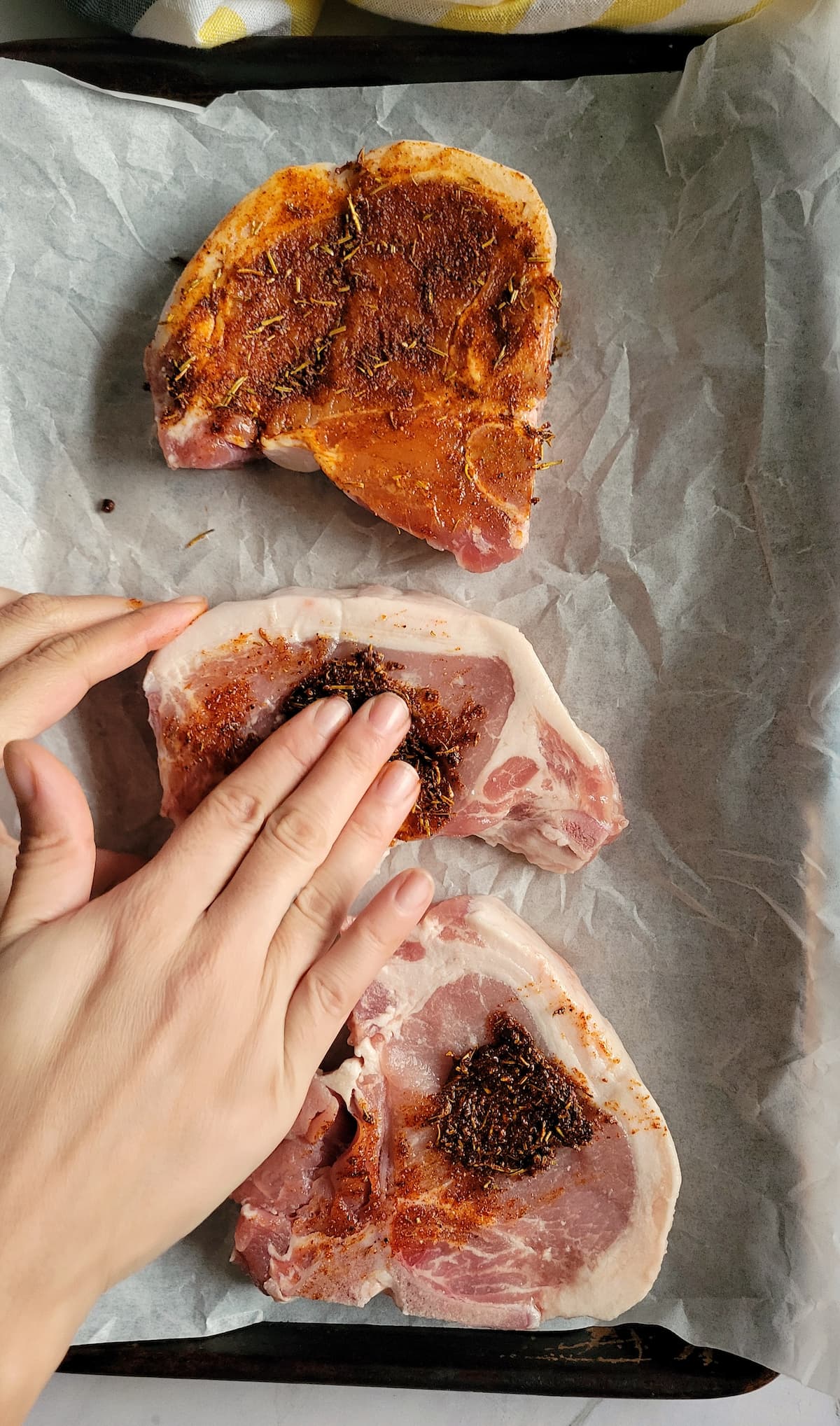 hand rubbing seasoning into 3 raw pork chops on a parchment lined baking sheet