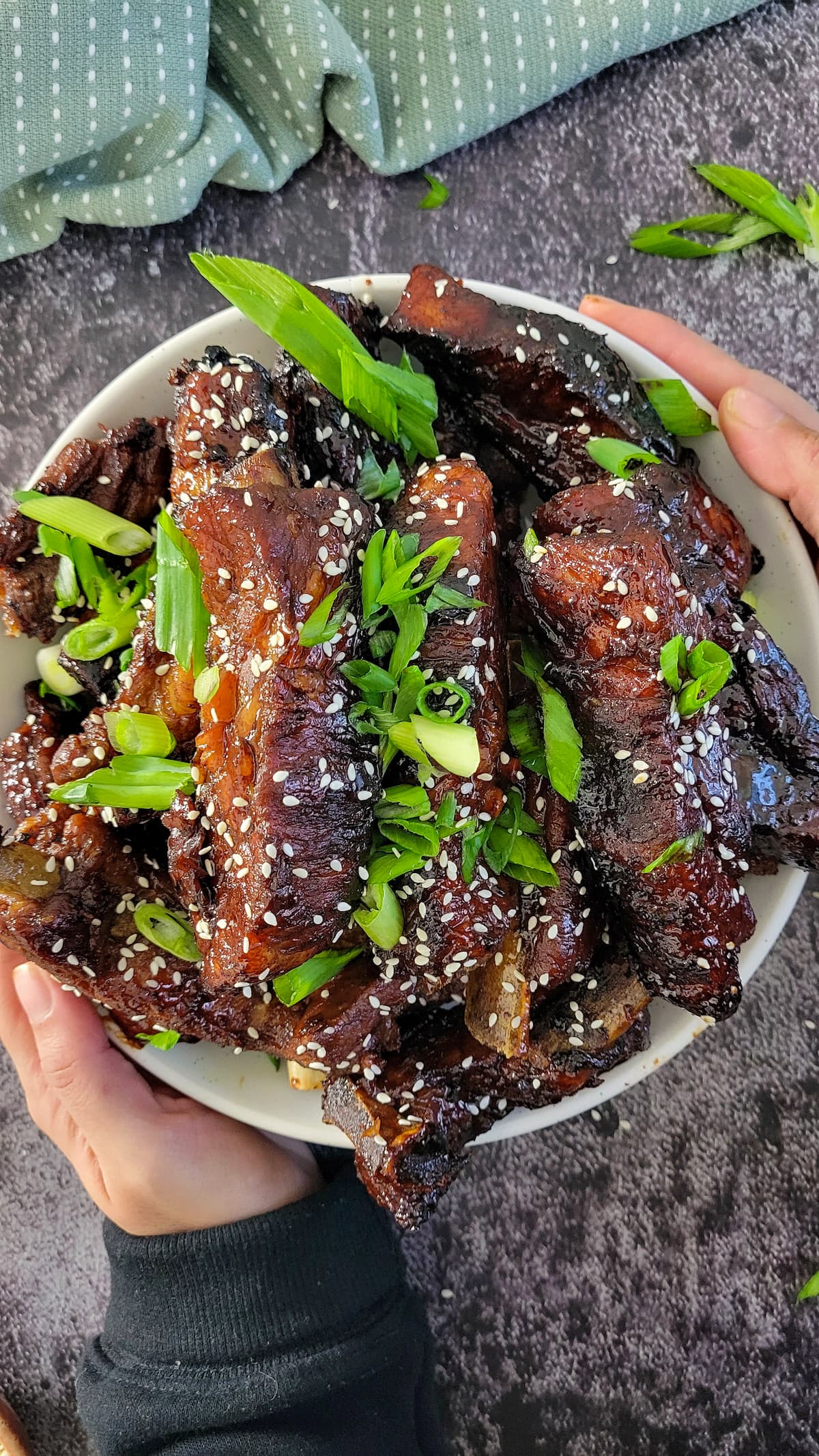hands holding a bowl of sticky asian ribs garnished with sesame seeds and green onions