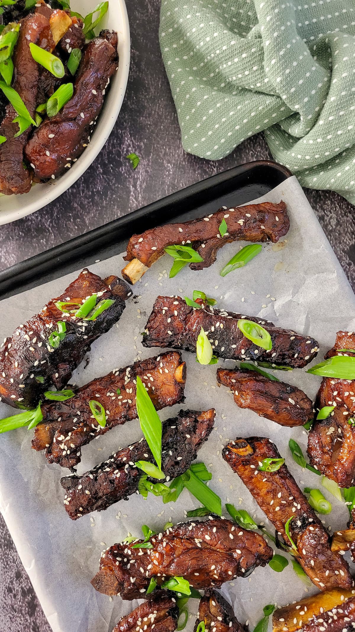 sticky asian ribs with green onions and sesame seeds on a parchment lined baking sheet, bowl with more in the background