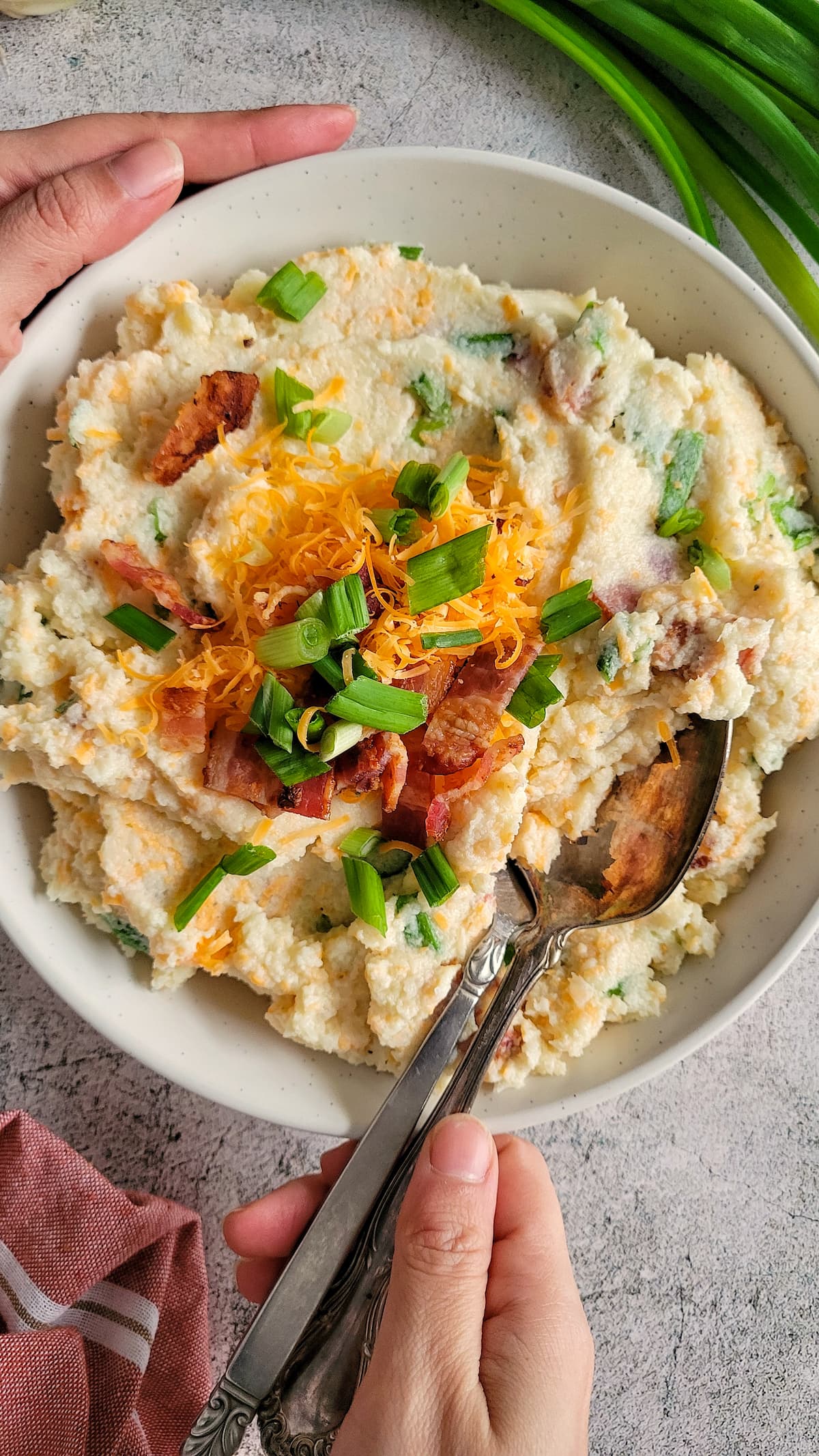 hands holding a bowl of mashed cauliflower with bacon, green onions and cheddar cheese