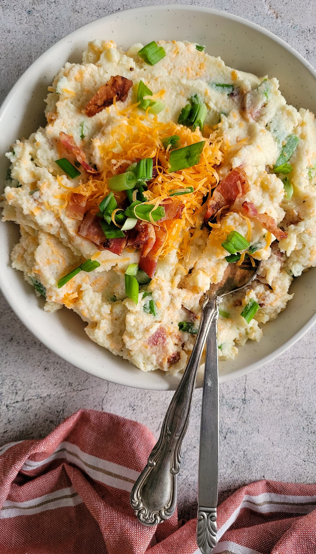 two spoons in a bowl of mashed cauliflower topped with cheddar, green onions and bacon