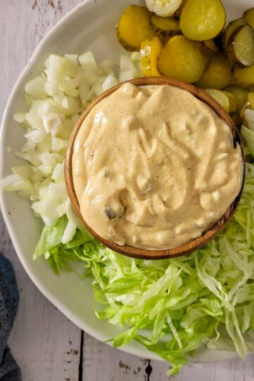 mac sauce in a bowl on a plate with sliced pickles, shredded lettuce and diced white onions