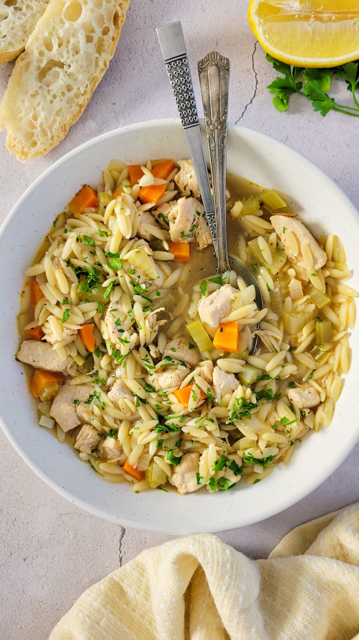 bowl of greek chicken soup with lemon, orzo, veggies and fresh chopped parsley, slices of bread and lemon in the background