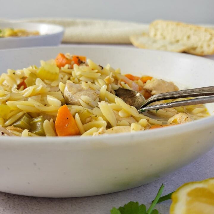 side view of a bowl of orzo chicken soup with carrots, lemon, parsley, bread and another bowl of soup in the background