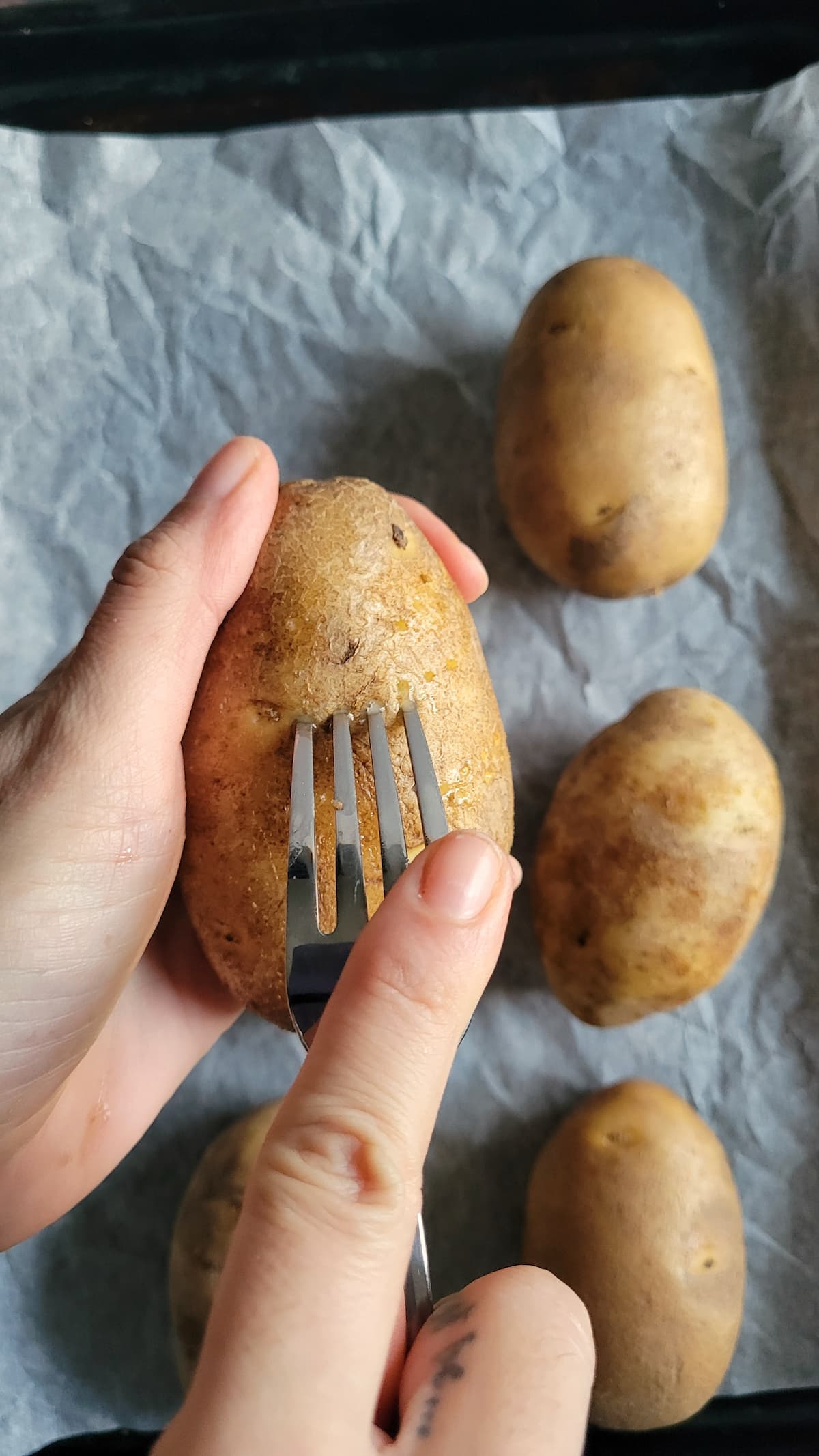 hand with a fork piercing a potato over a parchment lined baking sheet with more potatoes