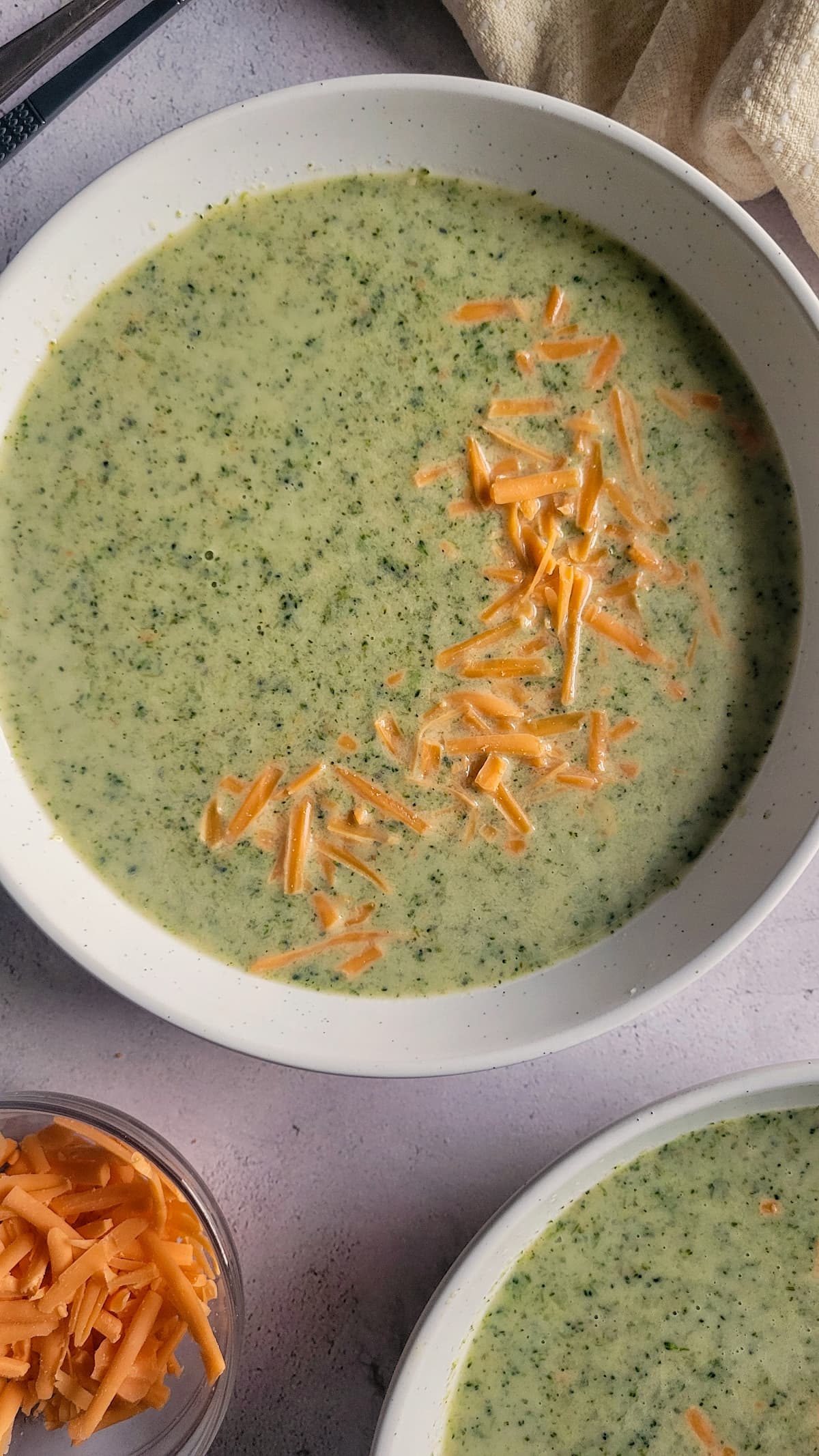 bowl of broccoli & cheese soup next to another bowl of it and a bowl of shredded cheddar