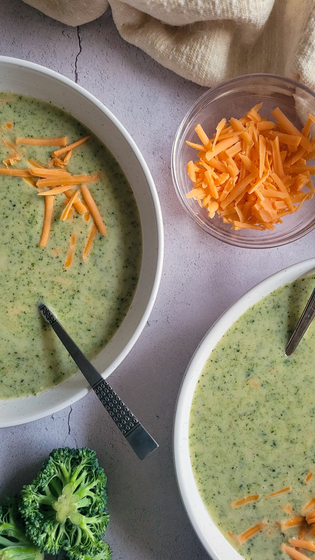 two bowls of broccoli & cheese soup next to some broccoli florets and a small bowl of grated cheddar cheese