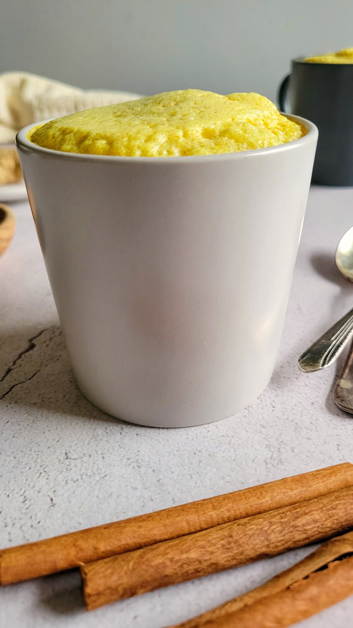 mug with yellow foam, cinnamon sticks in the front, another mug in the background