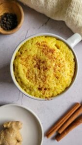 mug with turmeric milk topped with cinnamon next to black peppercorns, ginger and cinnamon sticks