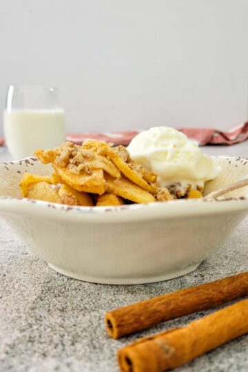 apple crisp in a bowl with ice cream, glass of milk in the background, cinnamon sticks in the front