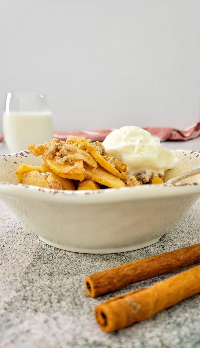 apple crisp in a bowl with ice cream, glass of milk in the background, cinnamon sticks in the front