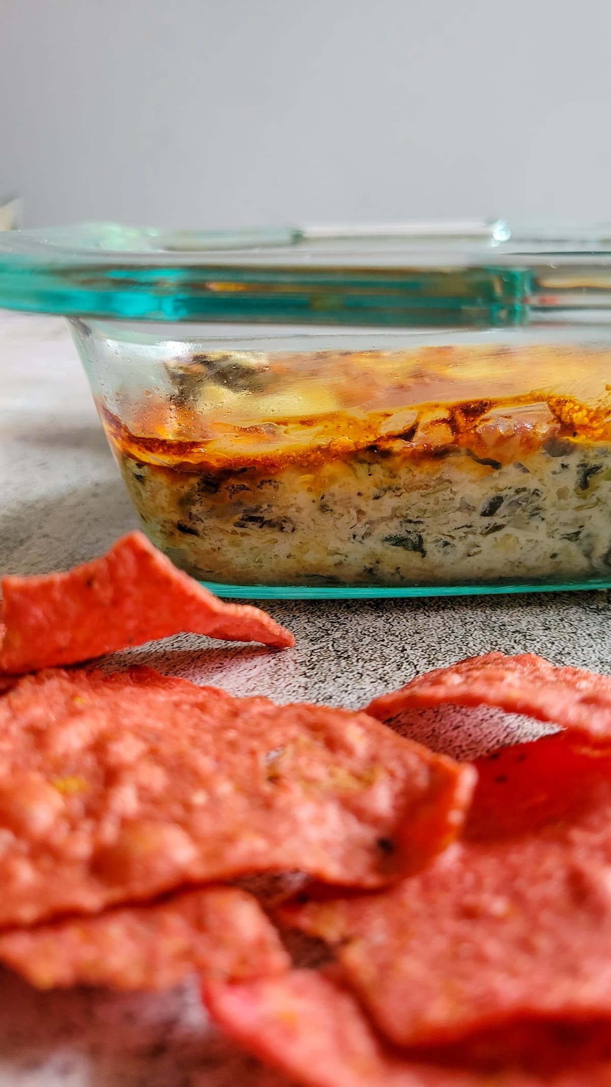 side view of spinach artichoke dip in a baking dish, red tortilla chips in the front