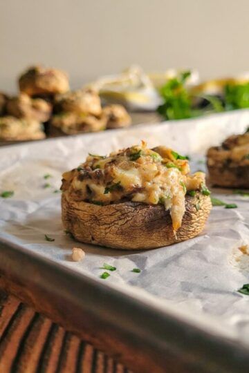 crab filled mushroom on a parchment lined baking sheet, plate stacked with more mushrooms in the background