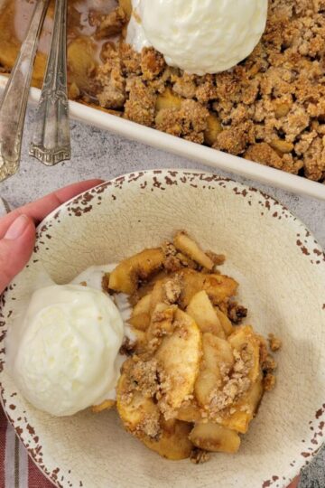 hands holding a bowl of apple crisp with ice cream, pan with more in the background