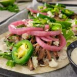 side view of brisket tacos with pickled onions, jalapenos, green onions and feta cheese on a parchment lined baking sheet