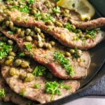 side view of veal scallopini with capers, parsley, and lemon in a cast iron skillet.