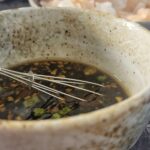 dumpling dipping sauce in a bowl with a whisk, plate of dumplings in the background